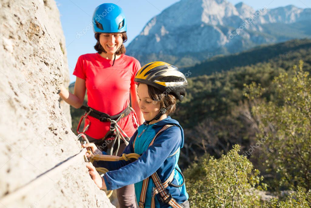 Mother teaches a child how to use safety equipment. A boy in a helmet goes through Via Ferrata. A woman instructs how to use a carabiner for belaying. Mountain tourism and mountaineering for children.