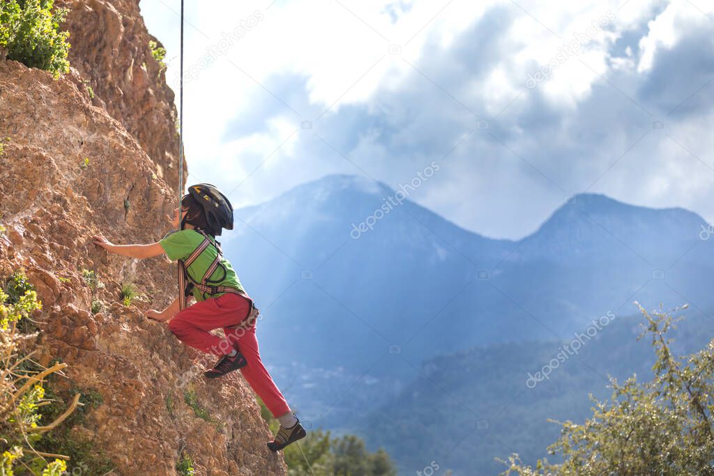 The child is climbing on a natural terrain. A boy climbs a rock on a background of mountains. Extreme hobby. Athletic kid trains to be strong. Rock climbing safety.