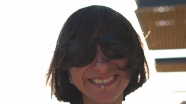 Piercingli ve güneş gözlüklü şehvetli bir kız güneşte poz verir ve rüzgarda çırpınan siyah saçlarına dokunur. Sakin