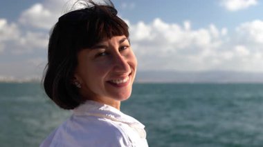 Çekici bir kadının yakın plan portresi, gülümseyen, esmer, kendine güvenen, deniz kenarında duran. Kız gülümser ve kameraya bakar.