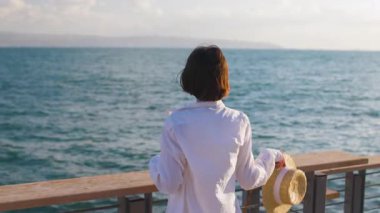 Sakin ve sakin bir kadın hasır bir şapka takıp okyanusa bakıyor. Deniz kenarında yaz tatili. Deniz kenarında özgürlük ve rahatlama.