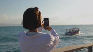 Güzel bir turist tatilde deniz manzarasından zevk alıyor ve cep telefonu kullanarak geminin fotoğraflarını çekiyor. sosyal içerik oluşturuluyor
