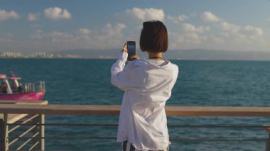 Güzel bir turist tatilde deniz manzarasından zevk alıyor ve cep telefonu kullanarak geminin fotoğraflarını çekiyor. sosyal içerik oluşturuluyor