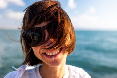 Piercingli ve güneş gözlüklü şehvetli bir kız, rüzgarda dalgalanan siyah saçlarıyla güneşte poz veriyor. Sakin esmer, altın akşam günbatımına karşı duruyor..