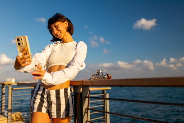 Kadın blogcu sosyal ağlar için içerik çekiyor. Gülümseyen turist, cep telefonuyla denizin arka planında selfie çekiyor. seyahat ve macera.