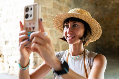 Neşeli, gülümseyen bir kadın eğleniyor ve Arap ülkesindeki eski bir şehrin sokaklarında fotoğraf çekiyor. Şık bir gezgin sosyal medya için simgelerin fotoğraflarını çekiyor. Telefonu olan kız..