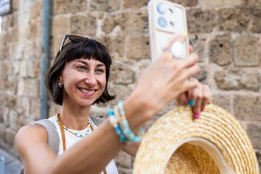 Neşeli, gülümseyen bir kadın eğleniyor ve Arap ülkesindeki eski bir şehrin sokaklarında fotoğraf çekiyor. Şık bir gezgin sosyal medya için simgelerin fotoğraflarını çekiyor. Telefonu olan kız..