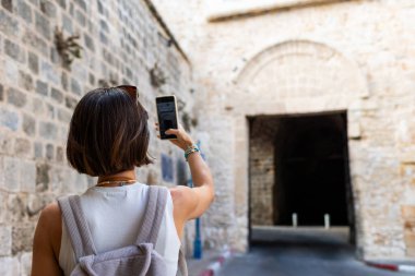 Neşeli, gülümseyen bir kadın eğleniyor ve Arap ülkesindeki eski bir şehrin sokaklarında fotoğraf çekiyor. Şık bir gezgin sosyal medya için simgelerin fotoğraflarını çekiyor. Telefonu olan kız..