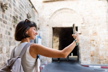 Neşeli, gülümseyen bir kadın eğleniyor ve Arap ülkesindeki eski bir şehrin sokaklarında fotoğraf çekiyor. Şık bir gezgin sosyal medya için simgelerin fotoğraflarını çekiyor. Telefonu olan kız..