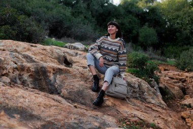 Sırt çantalı ve süveterli bir yürüyüşçü ormanda bir taşın üzerinde oturuyor.