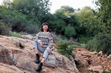 Sırt çantalı ve süveterli bir yürüyüşçü ormanda bir taşın üzerinde oturuyor.