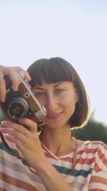Dikey çekim. Şık giyinmiş, odaklanmış bir esmer elinde fotoğraf makinesi tutuyor. Görüntü alıcısından bakıyor.
