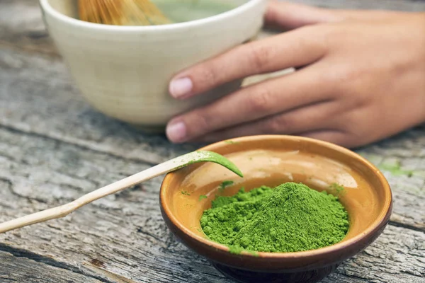 Matcha green tea powder in little clay bowl with girls hand holding a big bowl with a finished tea on the background