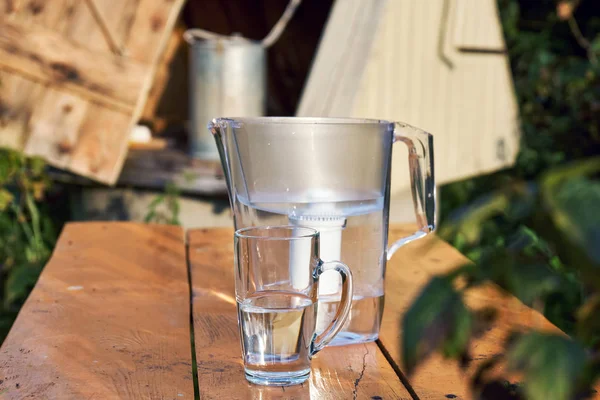 Water filter jug and a transparent cup of water standing on the wooden table with a village well on the background outdoors