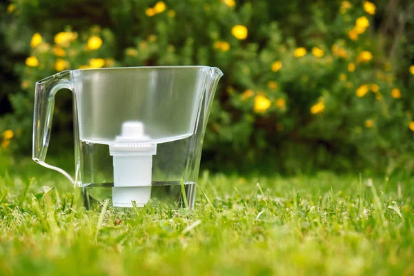 Filtre à eau debout sur l'herbe verte dans le jardin d'été avec des fleurs sur le fond — Photo