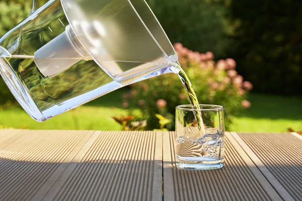 Pouring clear filtered water from a water filtration jug into a glass on the green summer garden background in a warm sunny summer day