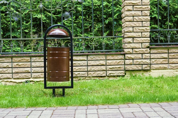 Metal trash bin on the green lawn near a brick fence of a private house in a russian cottage village