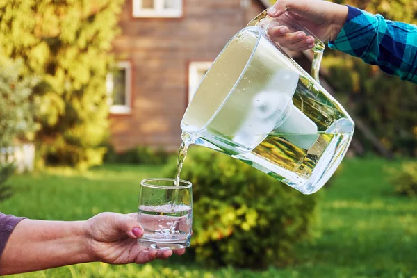 Girls hand pours clear filtered water from a water filtration jug into a glass which female hand holds in front of a country house in a warm summer evening