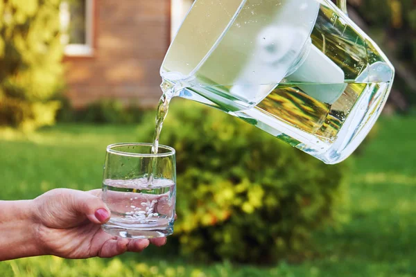 Pouring clear filtered water from a water filtration jug into a glass which female hand holding in front of country house in a warm sunny summer evening