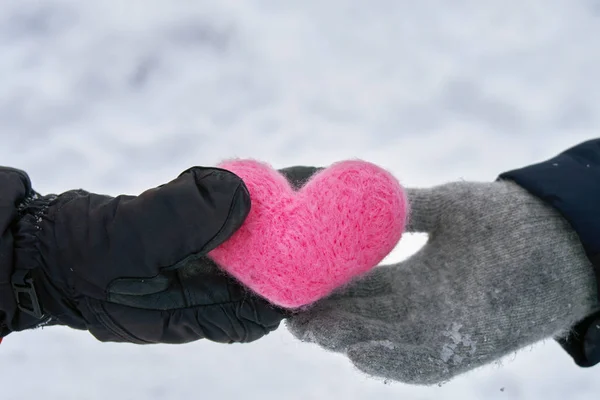 Male hand in black mitten gives a pink woolen heart to a female hand in woolen glove outdoors in winter. Love, helthcare, devotion,valentine's day concept.