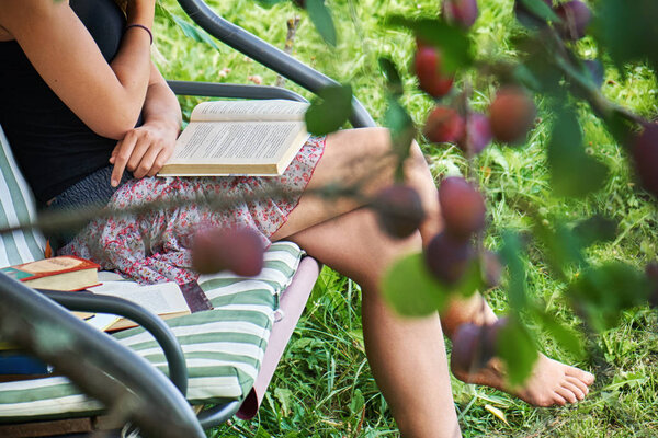 Teen girl reads a book sitting on a garden swing in summer garden.