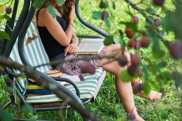Teen girl reads a book sitting on a garden swing in summer garden.