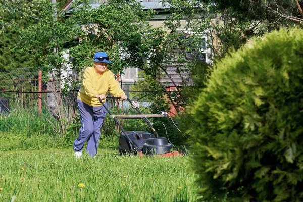 Mujer mayor corta el césped con una cortadora de césped eléctrica en el campo — Foto de Stock