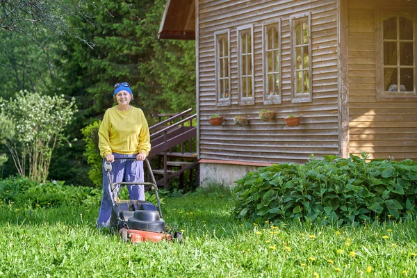 Mujer mayor sonriente corta el césped con una cortadora de césped eléctrica en el campo — Foto de Stock
