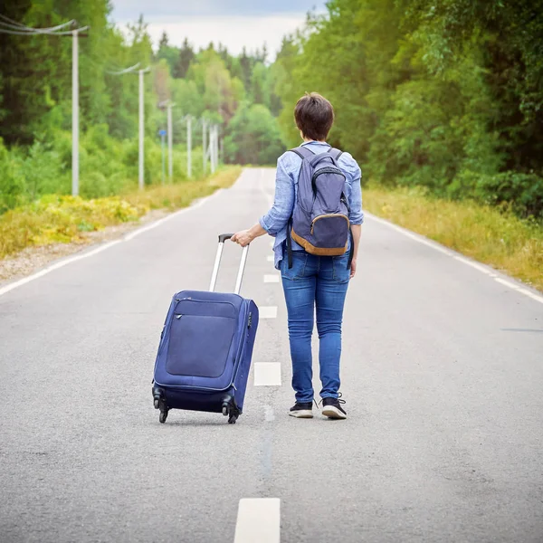 Turista mujer con una bolsa de viaje sobre ruedas y una mochila se aleja por la carretera — Foto de Stock