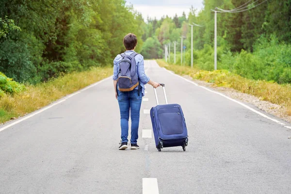 Turista mujer con una bolsa de viaje sobre ruedas y una mochila se aleja por la estrecha carretera — Foto de Stock