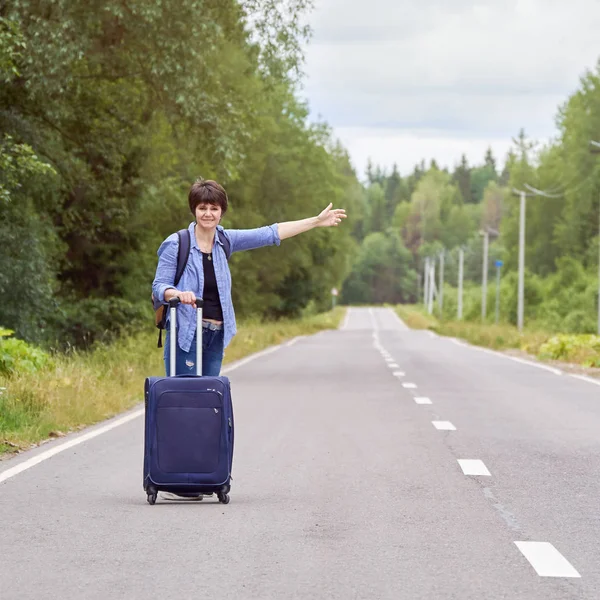 Nice middle-aged woman with a travel bag hitchhiking on the rural road in summer