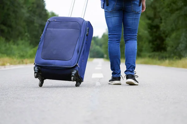 Piernas femeninas en jeans y zapatillas con una bolsa de viaje sobre ruedas se aleja por la carretera — Foto de Stock