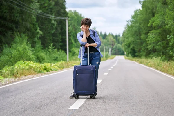 Sad woman with a luggage standing in the middle of a asphalt road and waiting for help