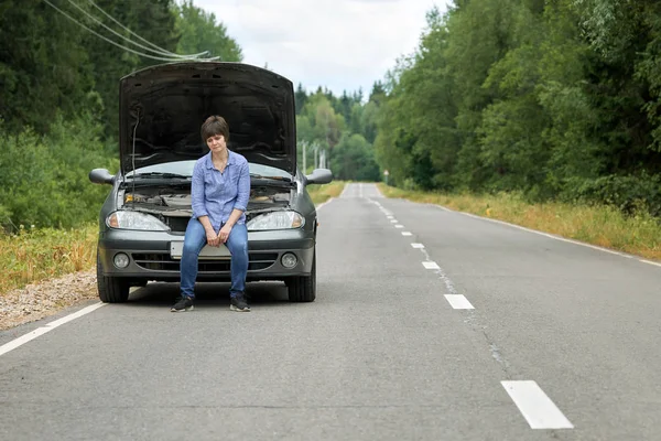 Upset woman sitting on the bumper of her old broken car on the road — Stock Photo, Image