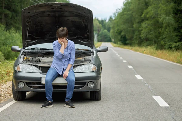 Mulher perturbada sentada no pára-choques de seu velho carro quebrado na estrada — Fotografia de Stock