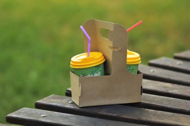 Closeup of cardboard coffee to go carrier, place for logo mockup, against sunlit summer park lawn on wooden bench clipart