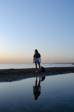 Sırt çantası olan bir kız sahilde duruyor ve gün batımını görüyor..