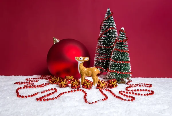 Christmas theme with miniature reindeer, decorated Christmas trees, presents and big bauble on red background