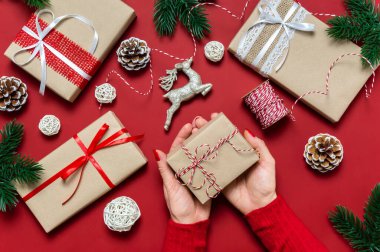 Kırmızı arka planda Noel hediyesini tutan kadın eller. Kadın eli, hediye kutuları, çam ağacı, takılar ve dekorasyonlu bayram kompozisyonu. Kış bayramı kutlaması. Üst manzara, düz uzanma.