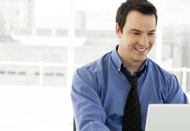 Mavi gömlekli genç bir işadamı ofiste dizüstü bilgisayarla çalışıyor.