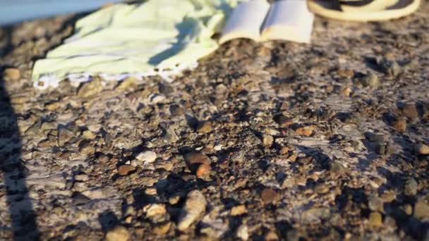 Un primo piano in movimento su uno scialle, cappello di paglia, libro aperto, spiaggia sabbiosa con onde blu. — Video Stock