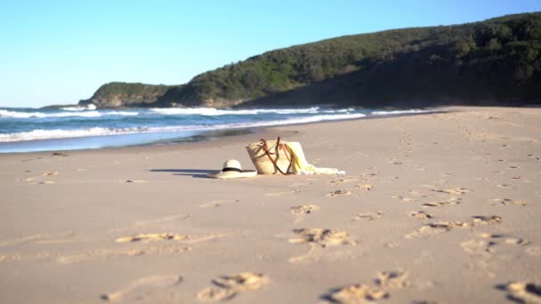 Napfényes tengerpart szalmakalapban, táskában és kendőben. Kék hullámok csapódnak a homokba.. — Stock videók