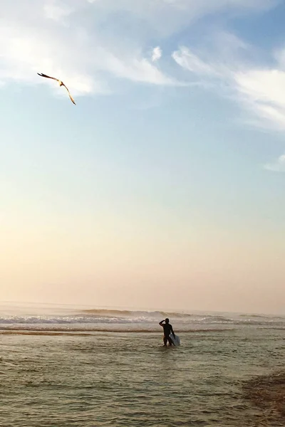 male surfer with surfboard standing in ocean at sunset