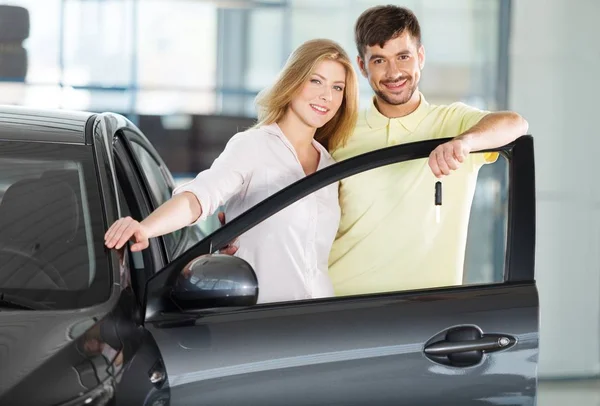 Feliz, joven pareja de pie junto al coche nuevo en la sala de exposición — Foto de Stock