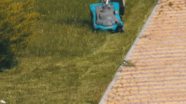 Человек с переносной электрической газонокосилкой косит зелёную траву на лужайке в парке — стоковое видео