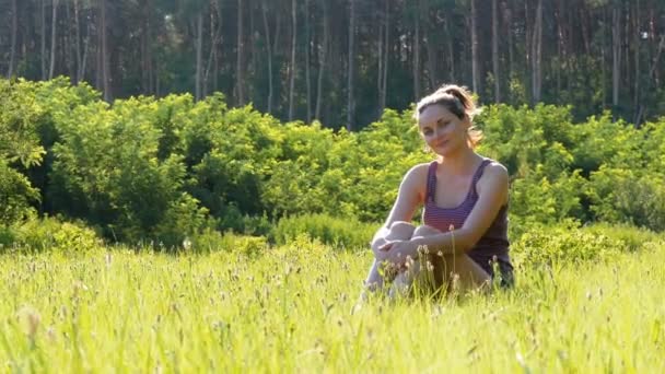 Glückliche junge Frau sitzt auf einem grünen Rasen in einem landschaftlich reizvollen Feld vor Sonnenuntergang. — Stockvideo