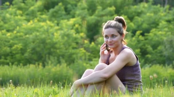 Glückliche junge Frau, die auf grünem Rasen sitzt und am Telefon oder Smartphone spricht — Stockvideo