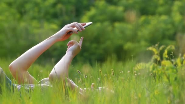 Glückliche junge Frau, die auf grünem Rasen liegt, nutzt Smartphone auf malerischem Feld bei Sonnenuntergang — Stockvideo