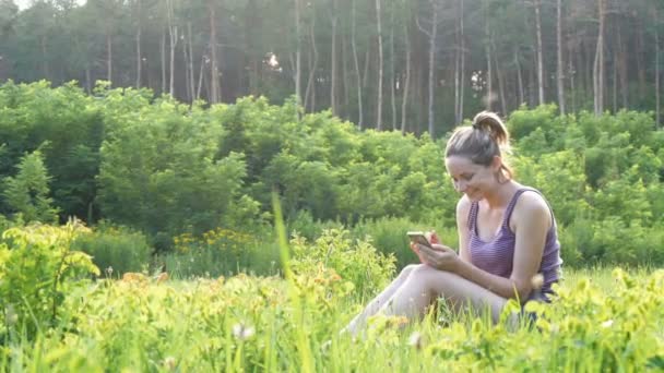 Joyeux jeune femme assise sur la pelouse verte et utilise un smartphone sur le terrain scénique au coucher du soleil Contexte — Video