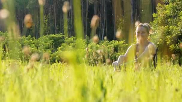 Mujer sonriente feliz se sienta en una hierba verde en un campo escénico sobre un fondo atardecer — Vídeo de stock